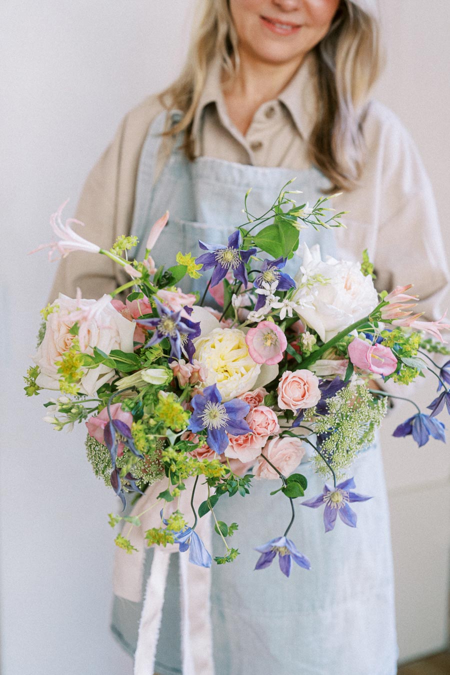 A florist wearing a beige shirt and blue apron holds a vibrant bouquet of pink roses, purple clematis, and various greenery, showcasing a fresh spring floral arrangement perfect for weddings and special occasions.