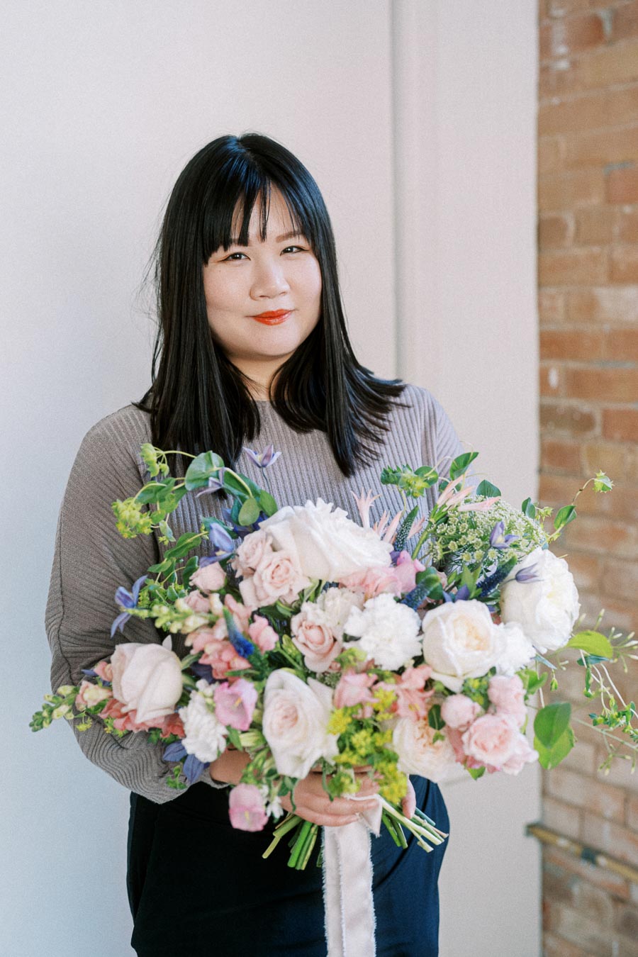 Woman holding a large, colorful bouquet of flowers with pink, white, and purple blossoms in a bright indoor setting next to a brick wall.