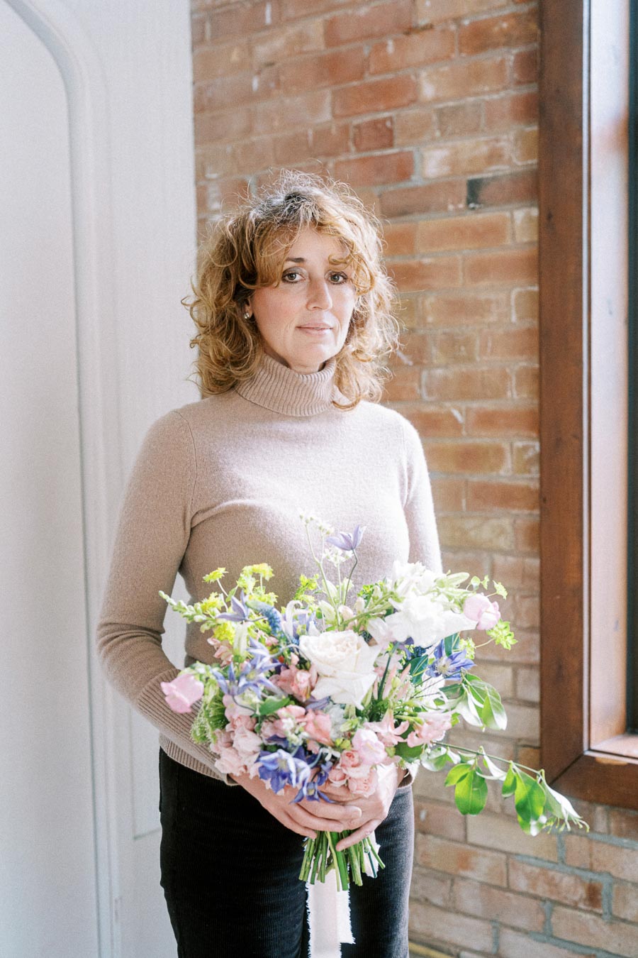 Woman holding a colorful bouquet of flowers indoors, standing by a brick wall in natural light, wearing a beige sweater.