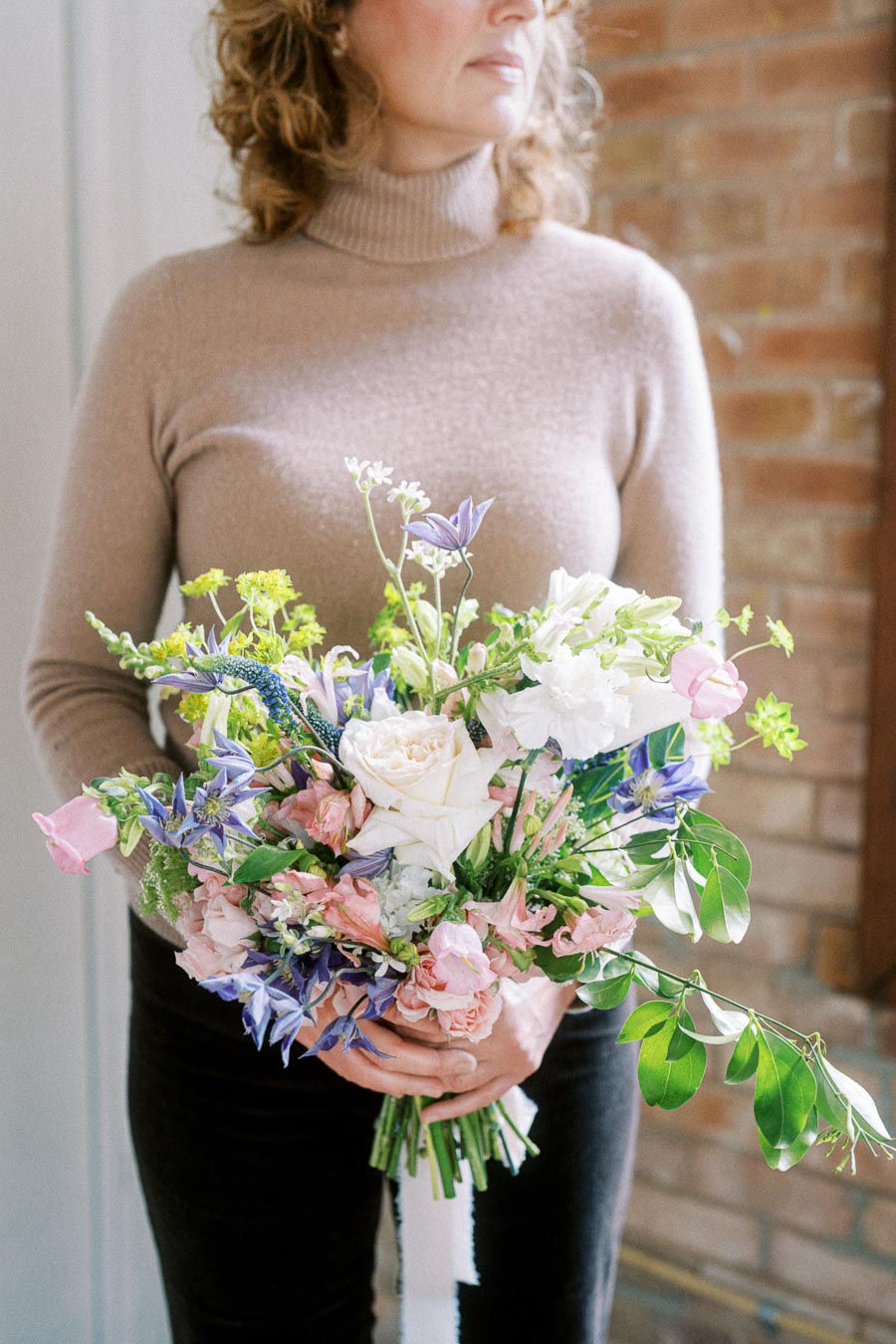 Woman holding a colorful floral bouquet with pink, white, and purple flowers in front of a brick wall, wearing a beige turtleneck sweater.