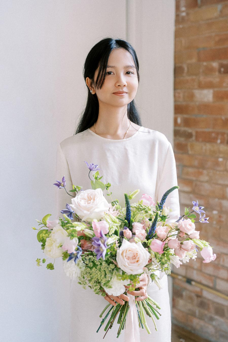 A woman holding a large bouquet of flowers with pink and purple blooms, wearing a cream-colored dress with a brick wall background.
