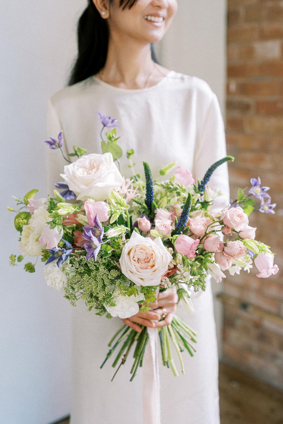 A woman in an elegant, light-colored dress holds a bouquet of pastel and white flowers featuring roses, accented with greenery, against a neutral background with a hint of brick wall.