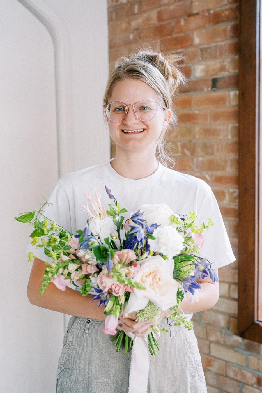 Smiling person holding a colorful bouquet of flowers in front of a brick wall background, wearing glasses and a casual white t-shirt.