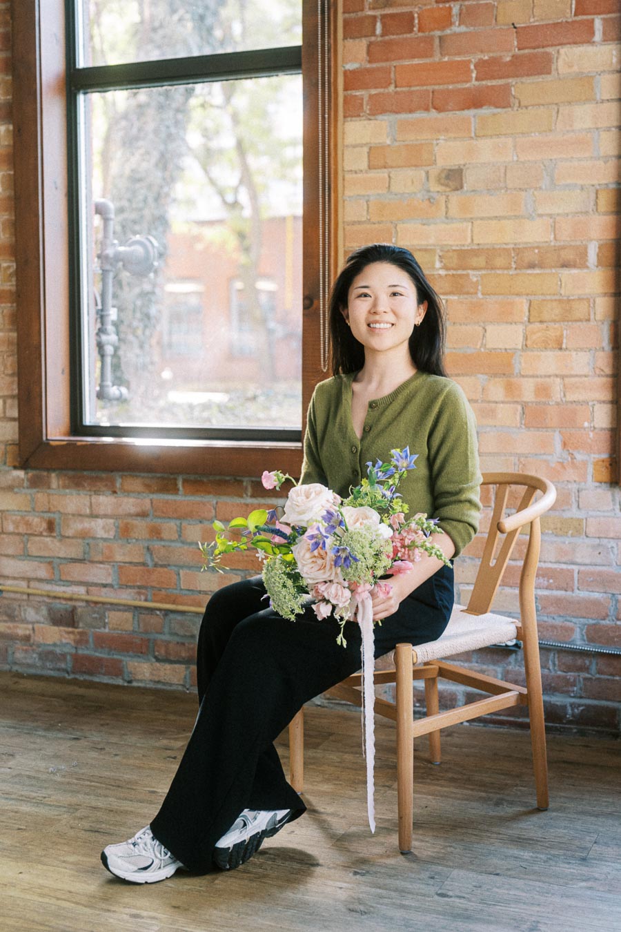 A person in a green sweater sits on a wooden chair holding a vibrant bouquet of flowers, set against a rustic brick wall with a large window and wooden floor, creating a serene indoor ambiance.