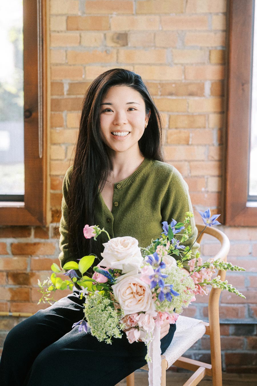 Smiling woman with long dark hair holding a vibrant bouquet of flowers, wearing a green sweater, seated indoors against a brick wall background.