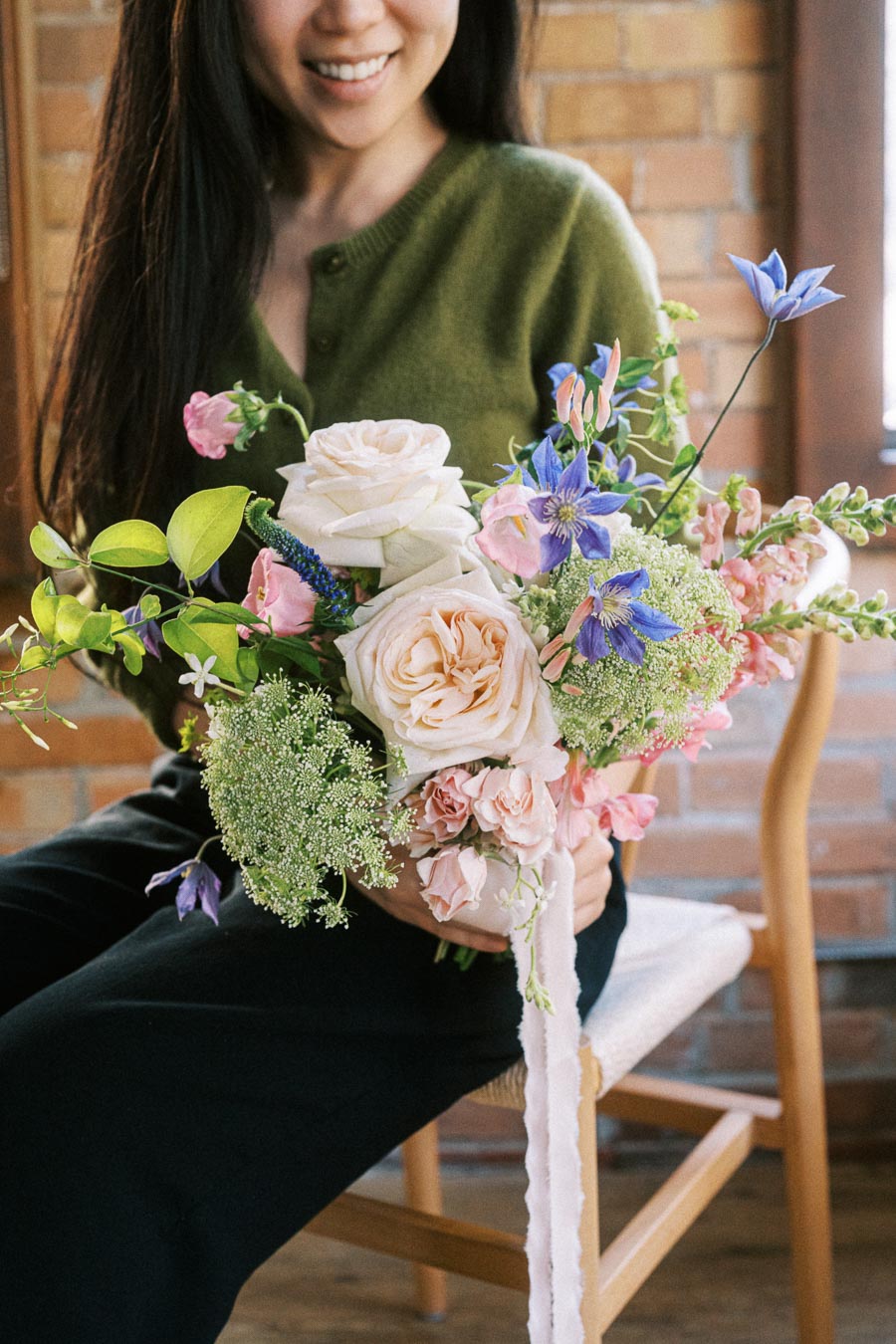A woman smiles while holding a colorful bouquet of flowers featuring pink roses, purple blooms, and lush greenery, sitting on a wooden chair against a brick wall background.