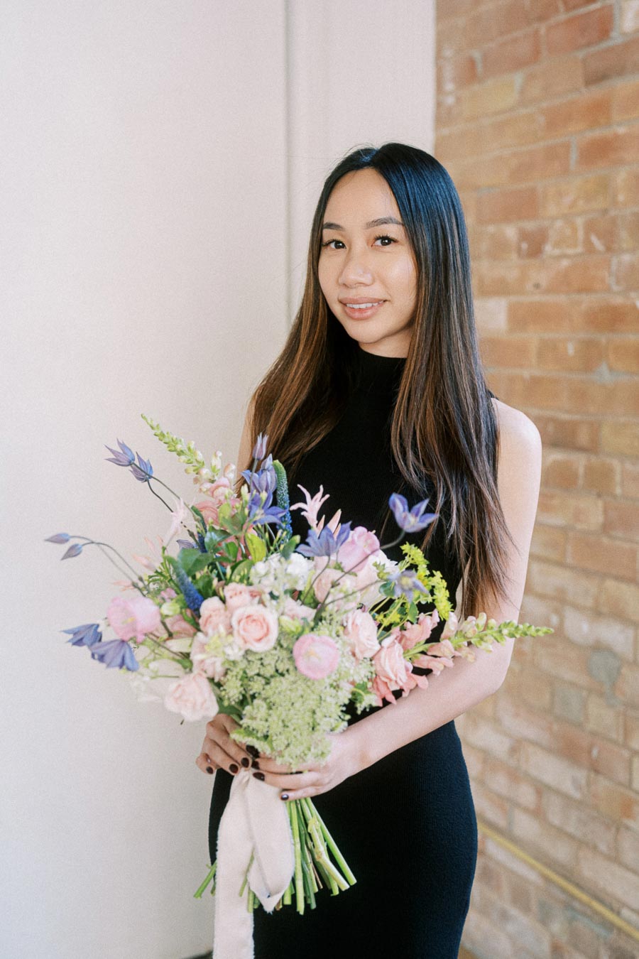 A person holding a colorful bouquet of flowers, featuring pink roses and purple blooms, wearing a black dress, in front of a brick wall background.
