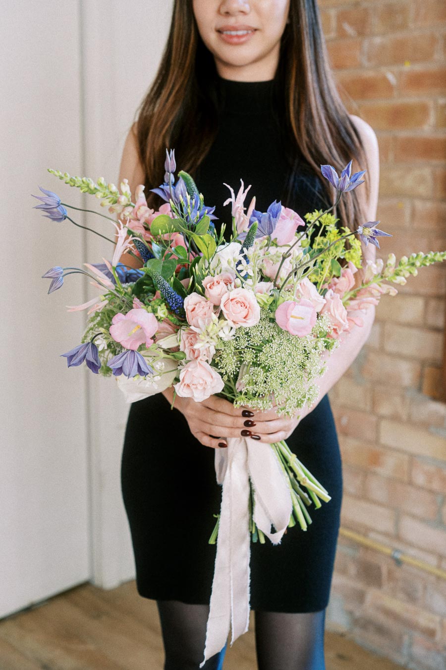 A woman in a black dress holding a vibrant bouquet of mixed flowers, including pink roses, blue bellflowers, and greenery, standing indoors against a brick wall background.