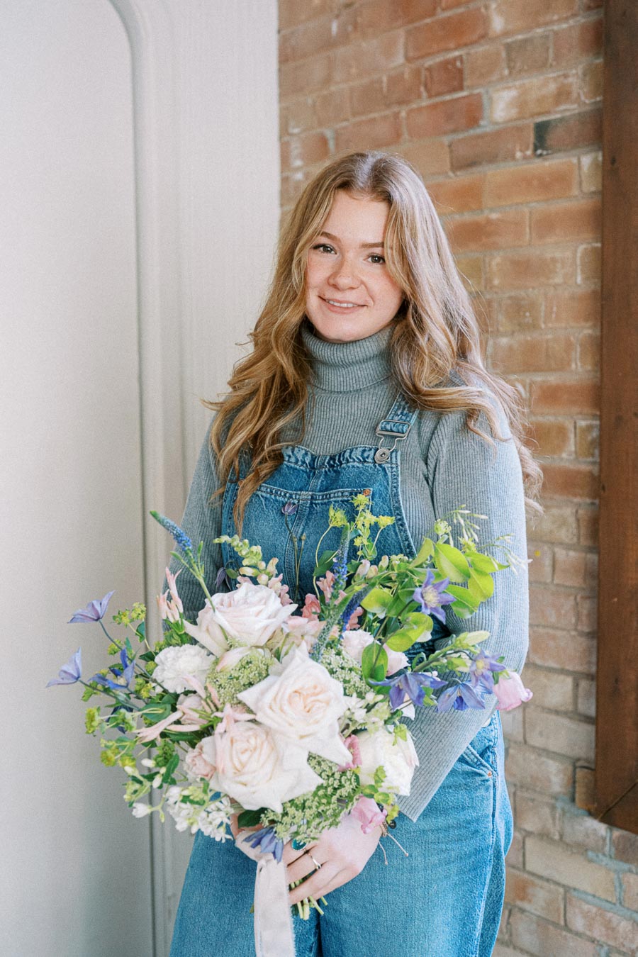 A person holding a bouquet of pink and white roses mixed with green foliage and purple flowers, standing against a brick wall background, wearing a gray turtleneck and denim overalls.