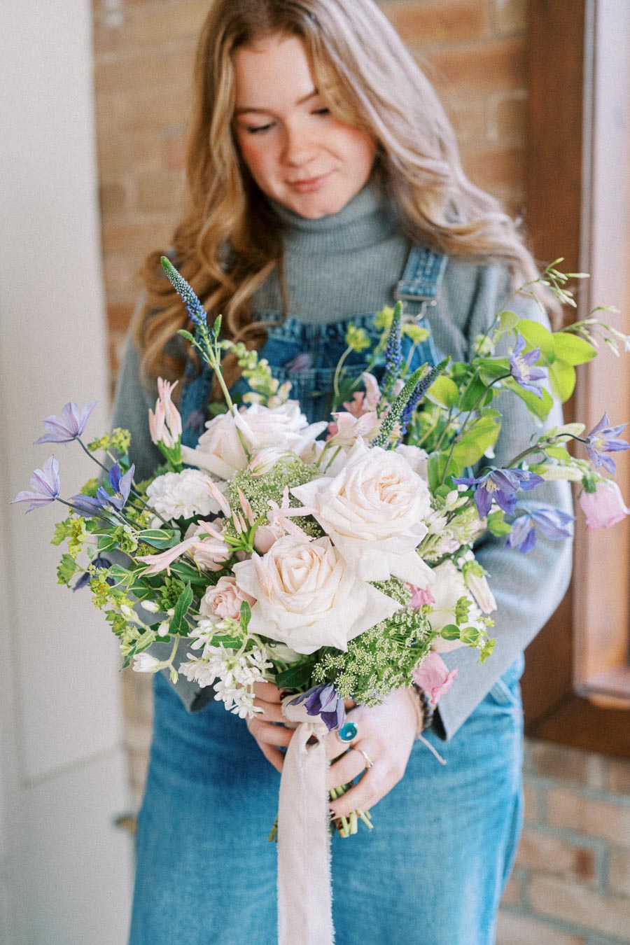 A woman in a gray sweater and denim overalls holding a bouquet of pink roses and assorted flowers, standing indoors with a blurred brick wall background.