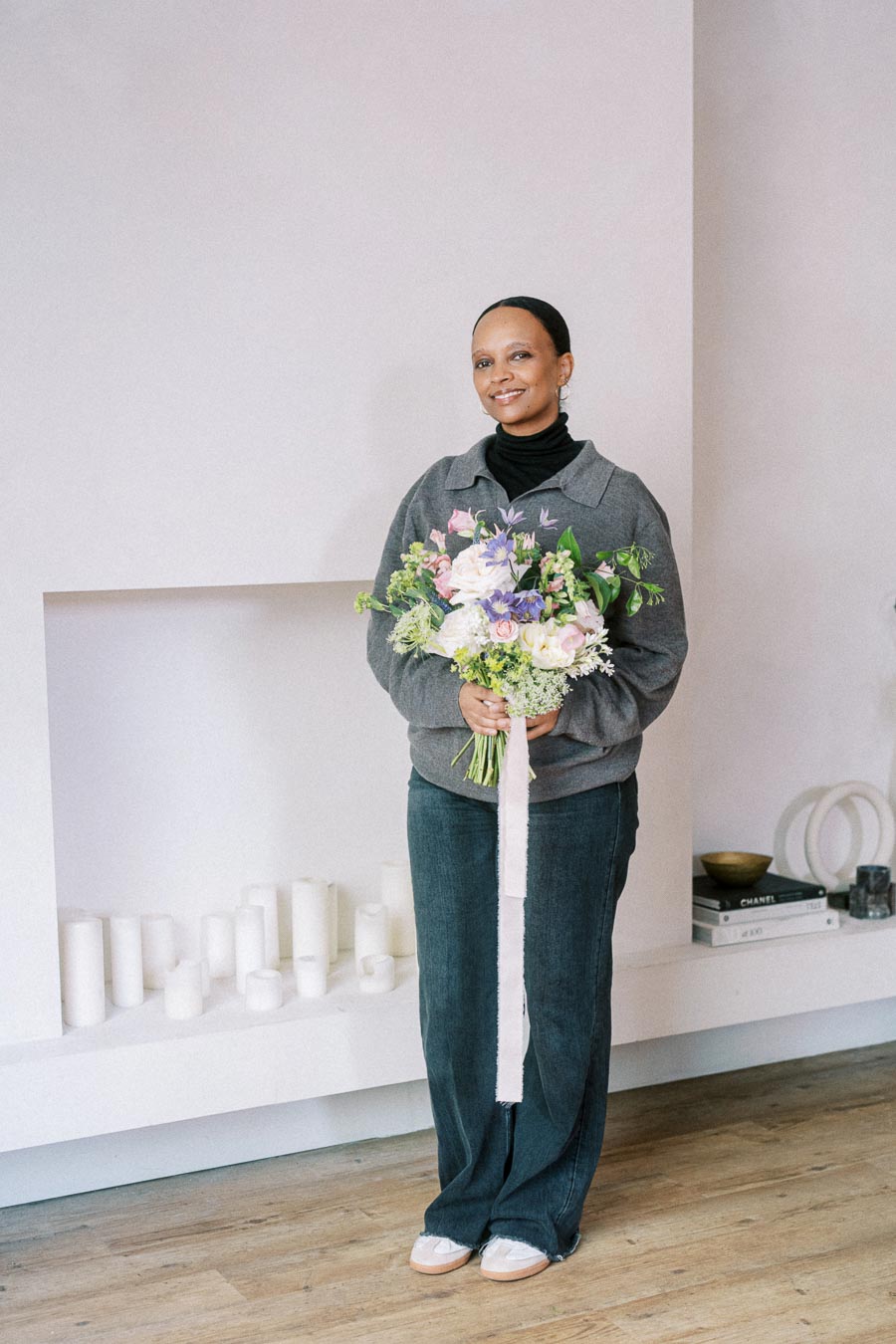 A person smiling while holding a colorful bouquet of flowers in a minimalist indoor setting with books and candles in the background.