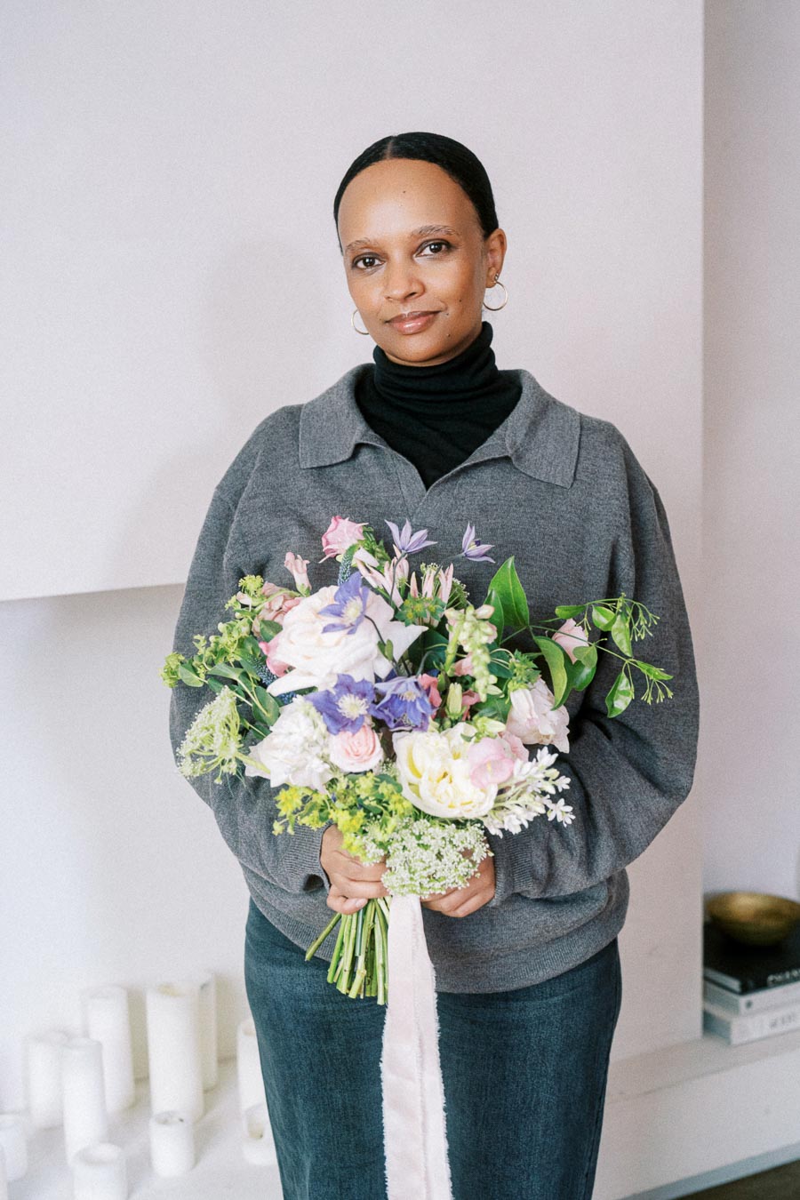 A person in a grey sweater holding a colorful bouquet of flowers, featuring pink, white, and purple blossoms, standing indoors with white candles in the background.