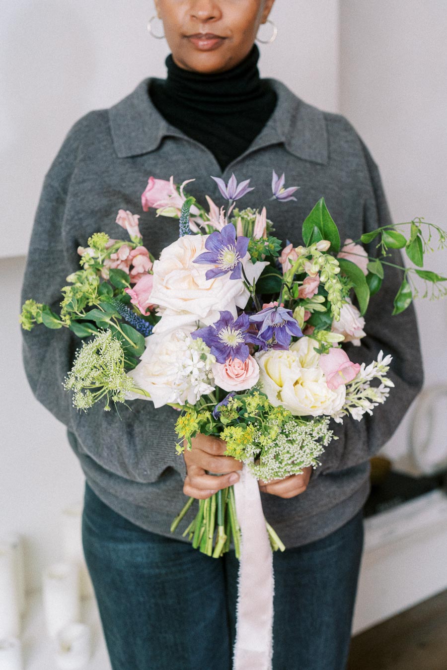 A person in a gray sweater holds a vibrant bouquet of mixed flowers, featuring white and pink roses, purple blooms, and lush greenery, creating an elegant and fresh floral arrangement.