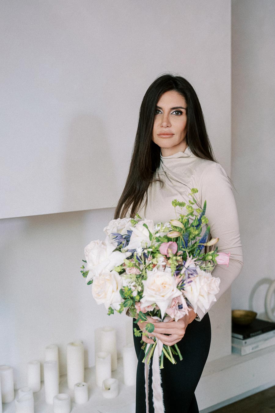Woman holding a large bouquet of white and pastel flowers in a modern, minimalist setting with white candles in the background.