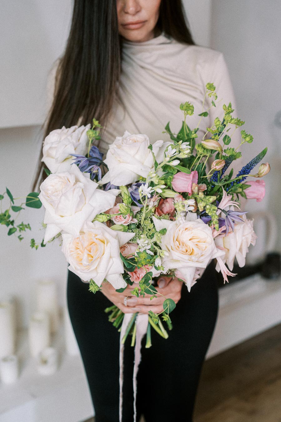 Woman holding a bouquet of white and pastel flowers, including roses and various greenery, in a cozy indoor setting.
