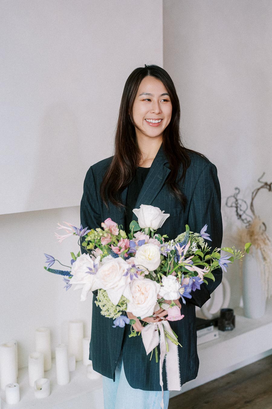 A smiling person holding a large bouquet of colorful flowers, standing in a bright room with minimalist decor.
