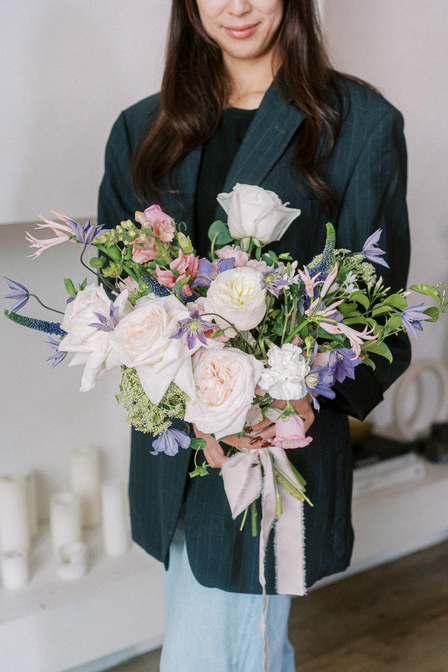 A person in a dark jacket holding a bouquet of mixed flowers with white, pink, and purple blooms, featuring roses and lilies, in a bright indoor setting.