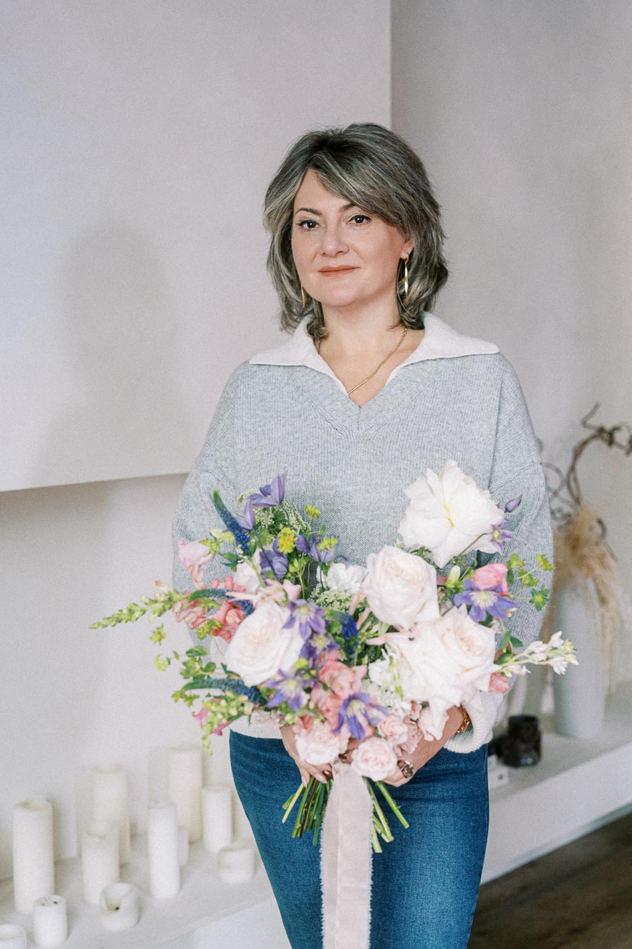 Woman holding a colorful bouquet of flowers, featuring pink roses and purple blooms, standing in a room with white candles in the background.