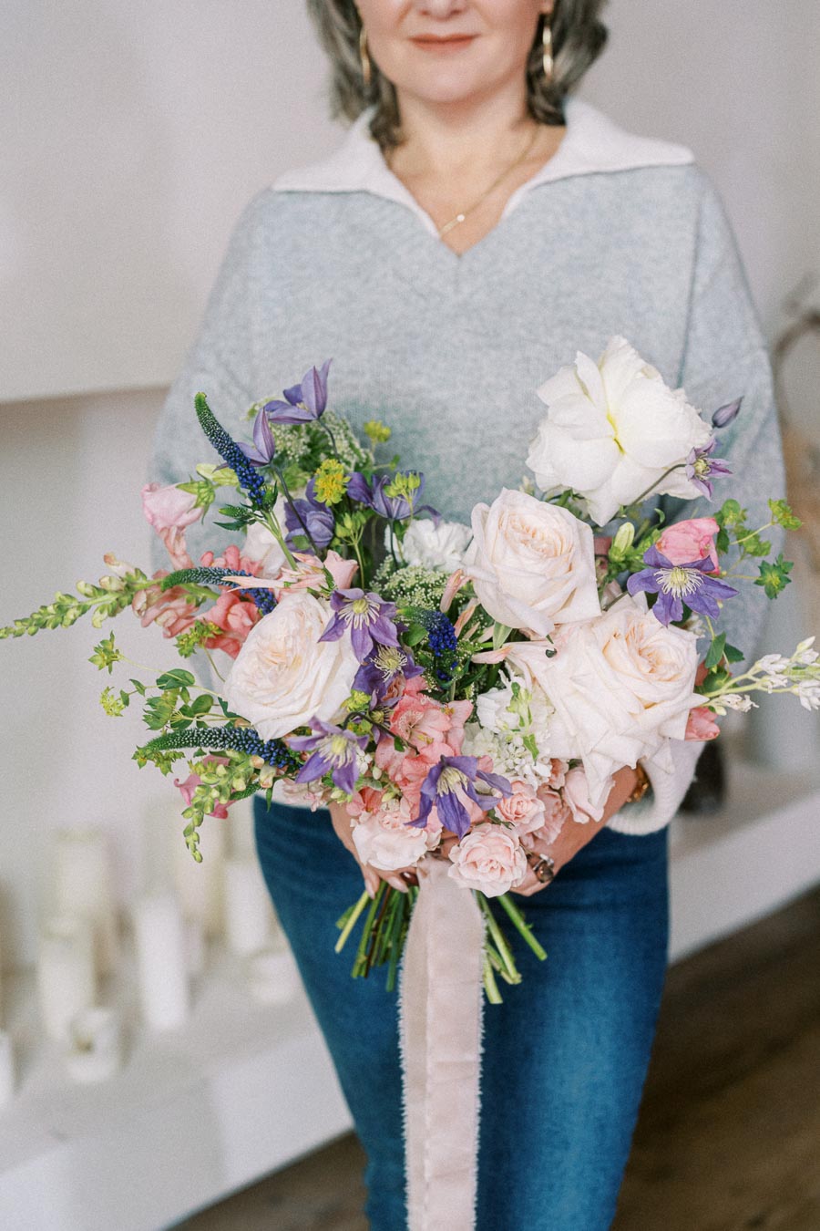 A woman in a blue sweater and jeans holding a vibrant bouquet of white, pink, and purple flowers with green foliage, in a bright indoor setting.