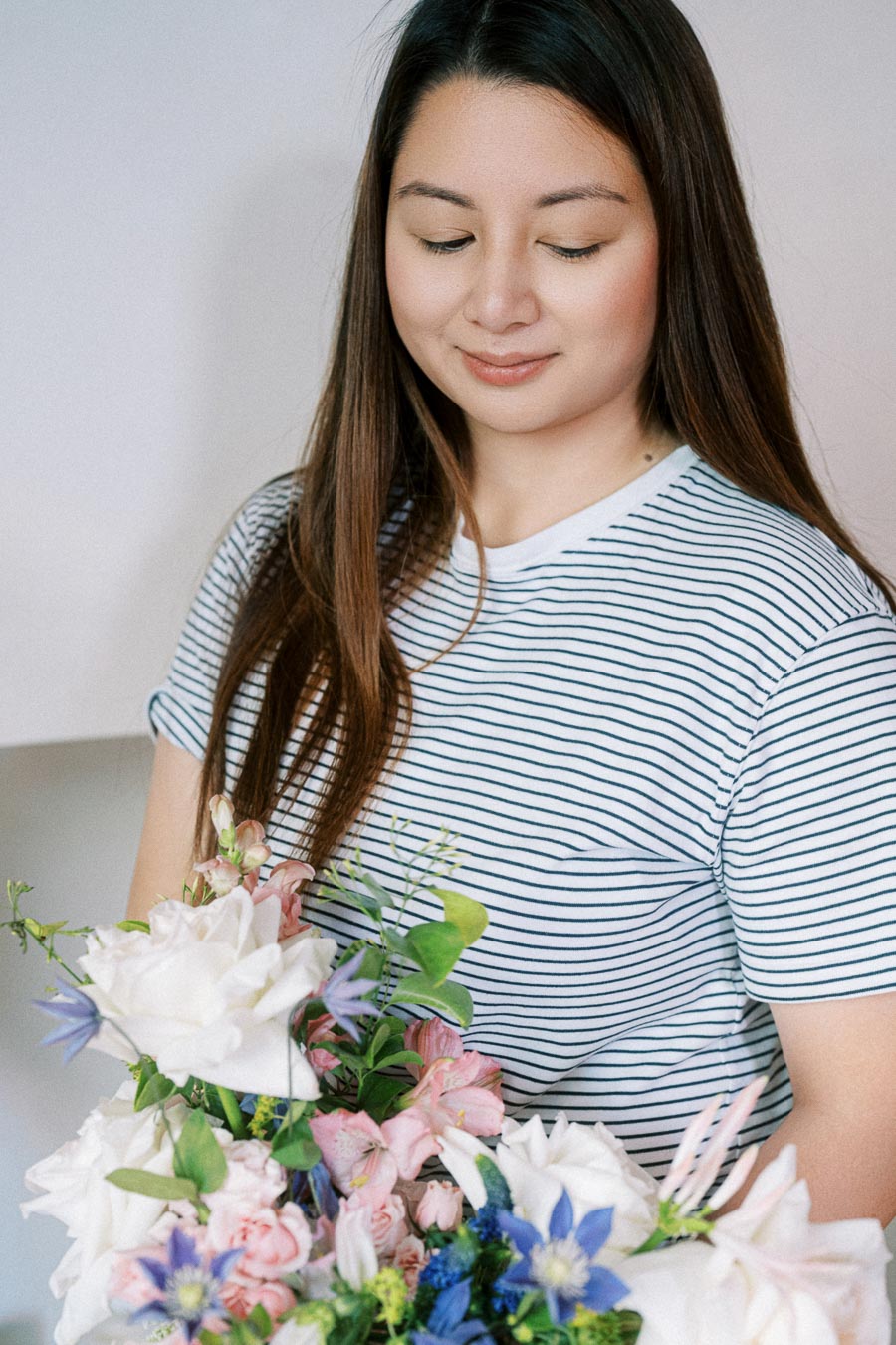 A woman in a striped shirt smiles gently while holding a vibrant bouquet of pink, blue, and white flowers.