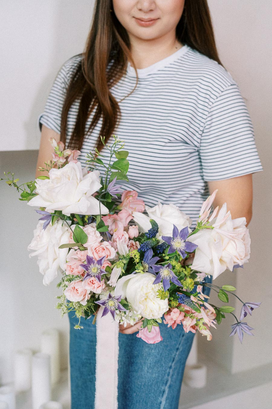 Woman holding a vibrant bouquet of mixed flowers, featuring pink roses, white peonies, and purple blooms, against a neutral background.
