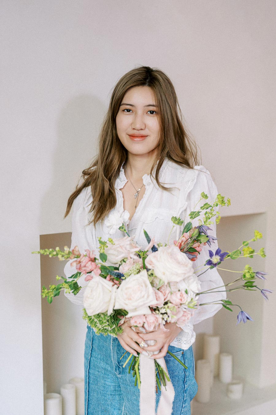 Young woman holding a bouquet of pink and white flowers, wearing a white blouse and blue jeans, smiling against a neutral background.