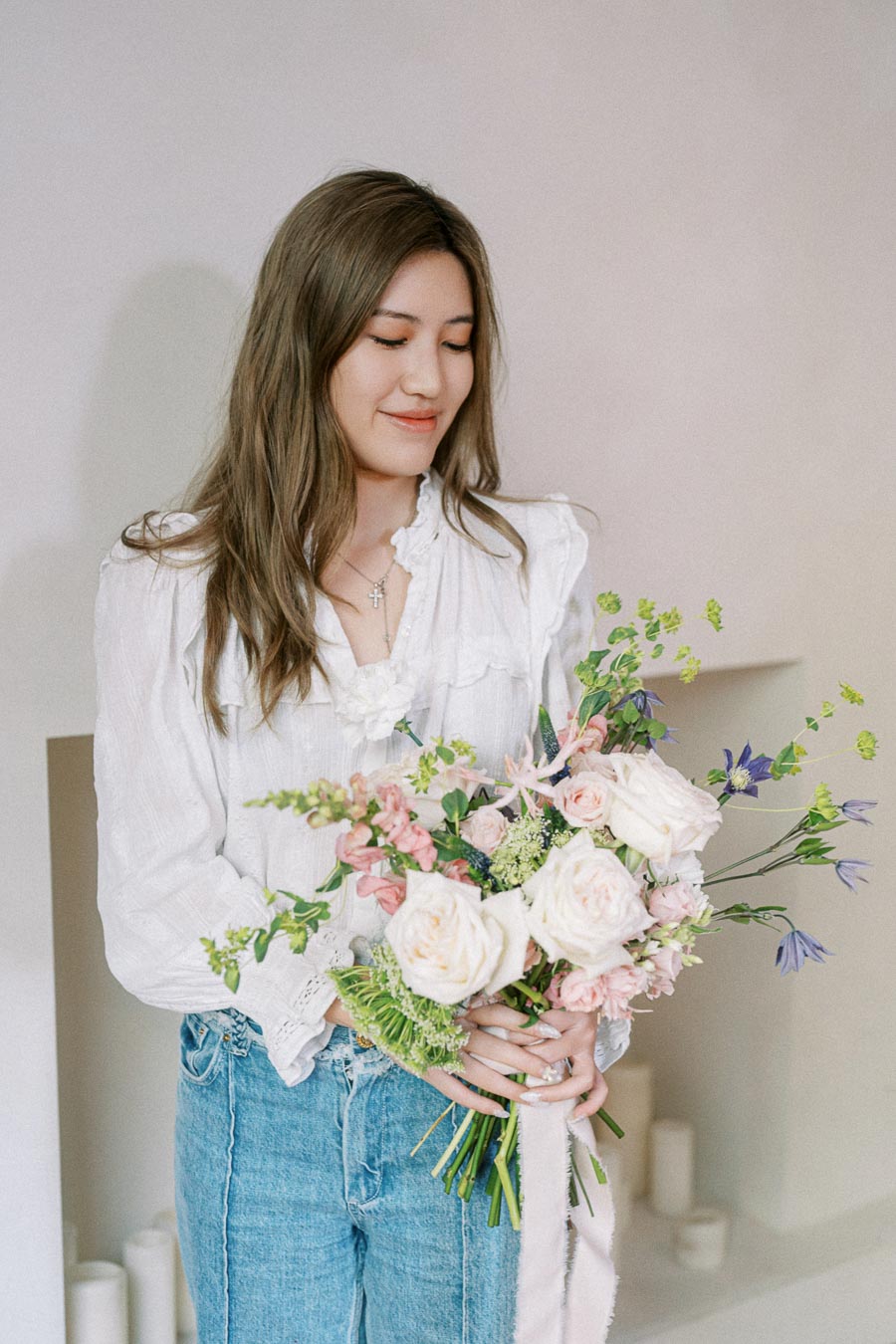 Young woman holding a bouquet of white and pink flowers, wearing a white blouse and jeans, standing against a light background.