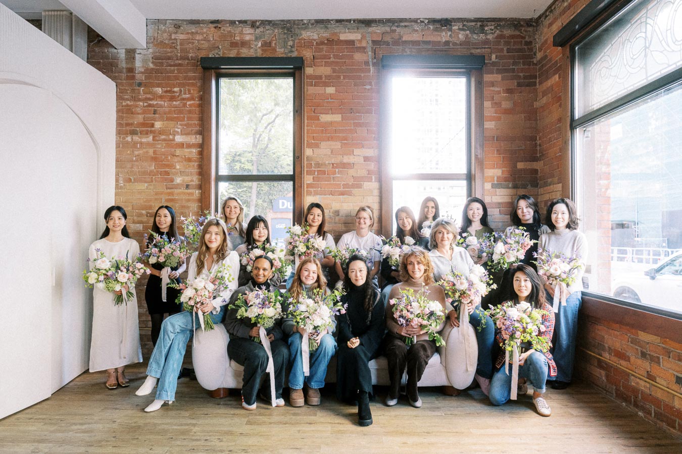 Group of people posing with floral arrangements, sitting and standing in a bright room with brick walls and large windows.