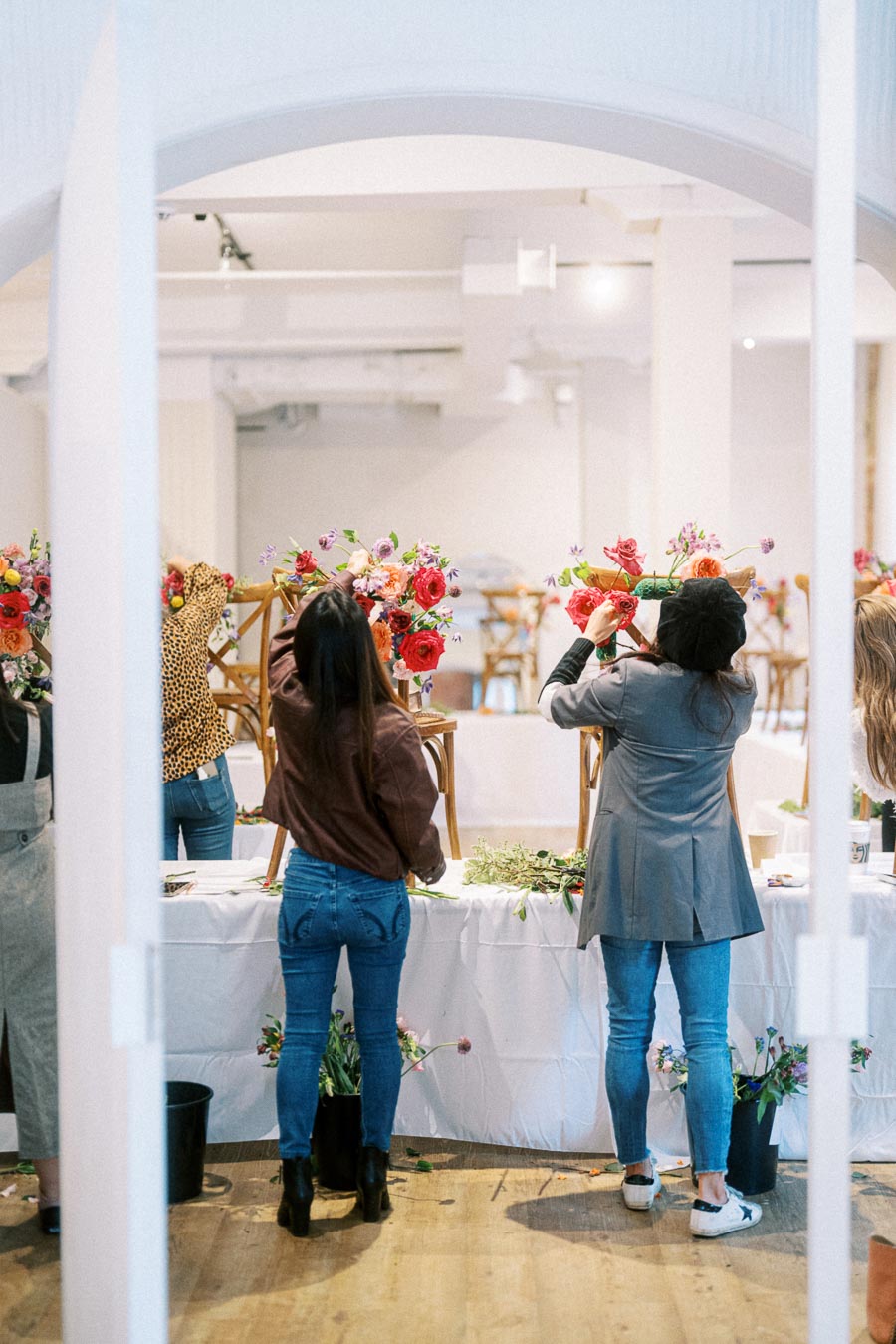Group of people arranging colorful floral decorations on wooden chairs at an indoor workshop, showcasing flower arrangement skills and creativity.
