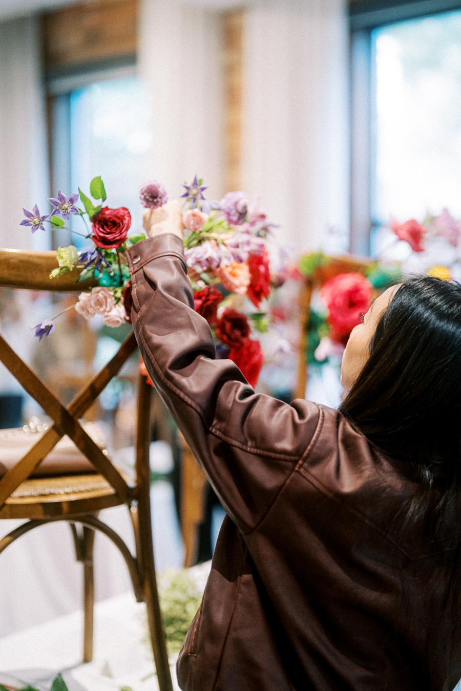 A person arranging vibrant floral decorations on a wooden chair during an indoor event setup, showcasing colorful flowers and elegant design.