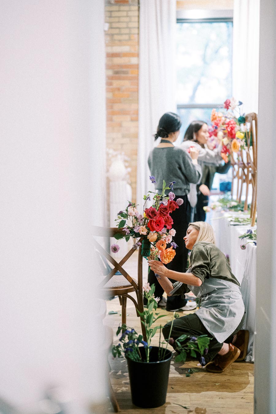 Florists arranging vibrant flowers for a decorative display in a sunlit room with a rustic brick wall, highlighting event preparation and floral artistry.