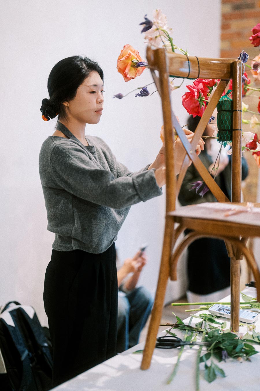 Florist arranging colorful flowers on a wooden chair, showcasing creative floral design with focus on vibrant roses and greenery in a workshop setting.