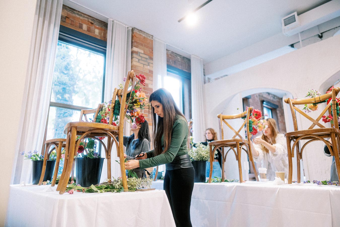Women creating floral arrangements on wooden chairs set on tables in a bright, airy room with large windows.