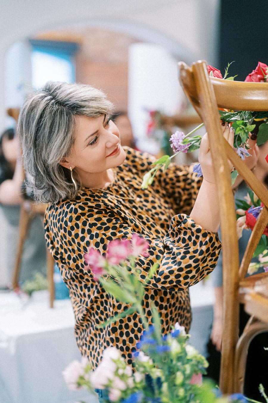 A woman in a leopard print top arranging vibrant flowers on a wooden chair, focusing intently in a creative workshop setting.