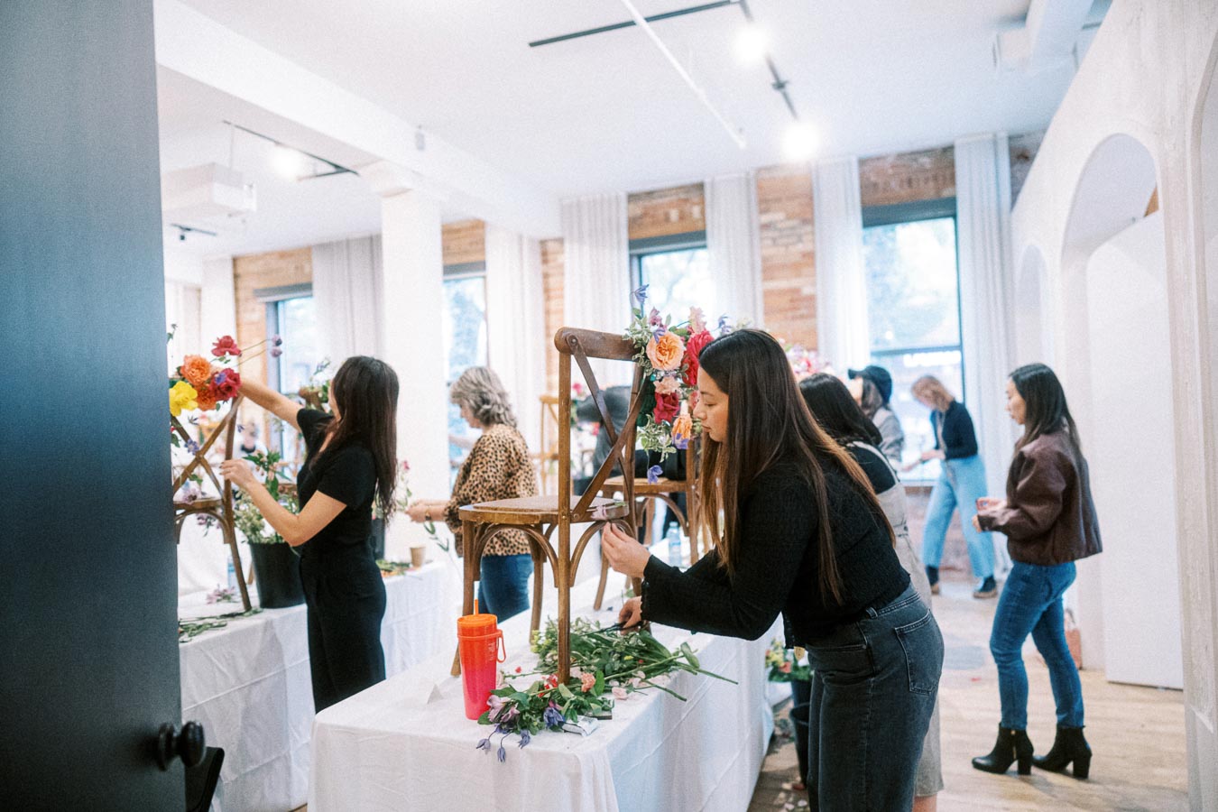 Women participating in a floral arrangement workshop, decorating wooden chairs with colorful blooms in a bright, modern studio space.