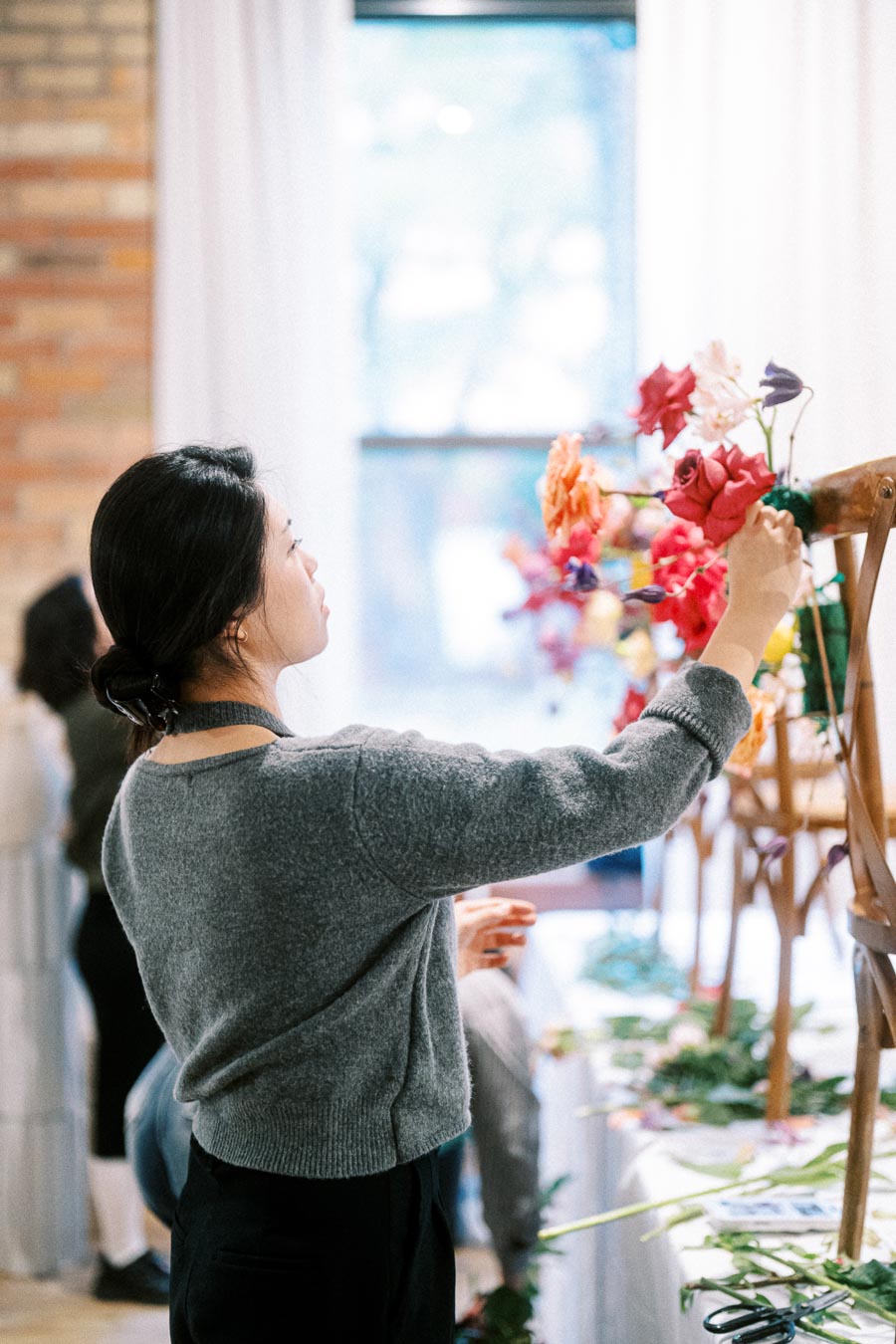 A woman in a grey sweater arranging colorful flowers during a floral design workshop indoors.