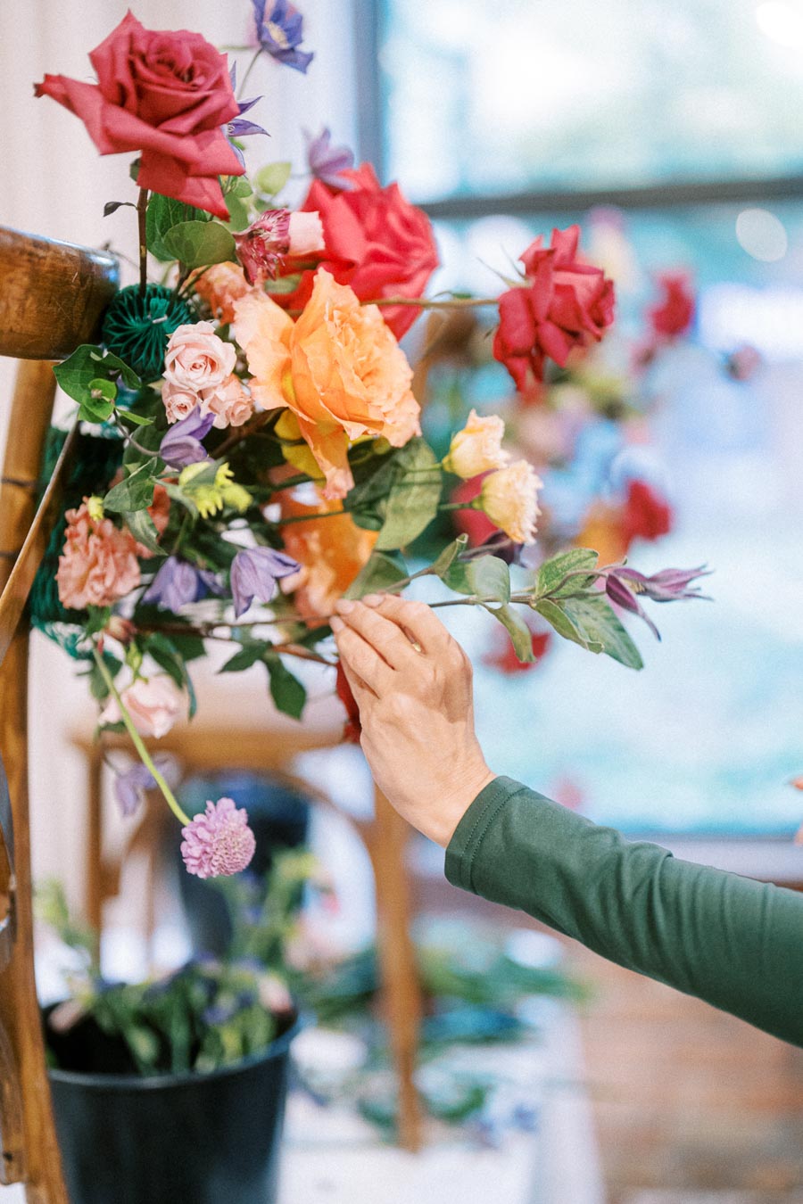 A person arranging a vibrant floral display featuring red and orange roses, pink and purple blossoms, and green leaves on a wooden structure indoors.
