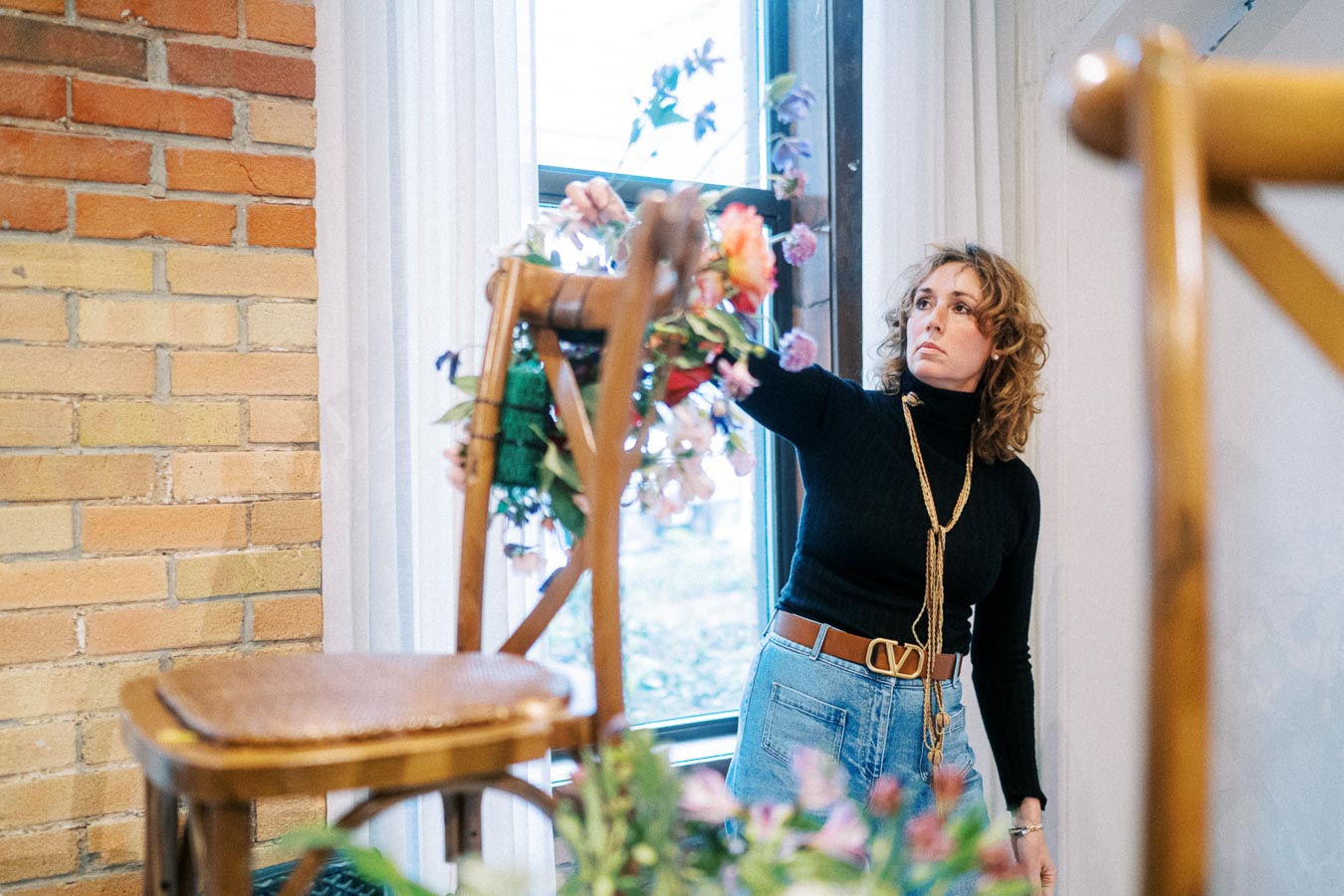 Woman arranging floral decorations on a wooden chair inside a bright room with brick walls and large windows.