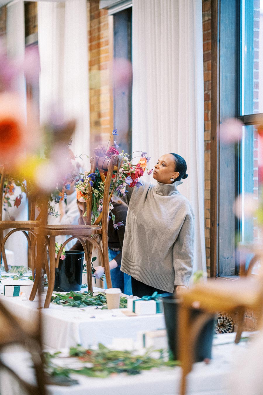 A person arranging colorful flowers on an upside-down chair at a wedding or event venue, surrounded by decor materials on a table, wearing a thick sweater, with a bright and airy background showcasing a brick wall and window.