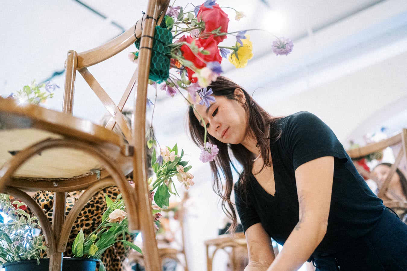 A woman arranging a creative floral display on a wooden chair, using a variety of colorful flowers in a modern indoor setting.