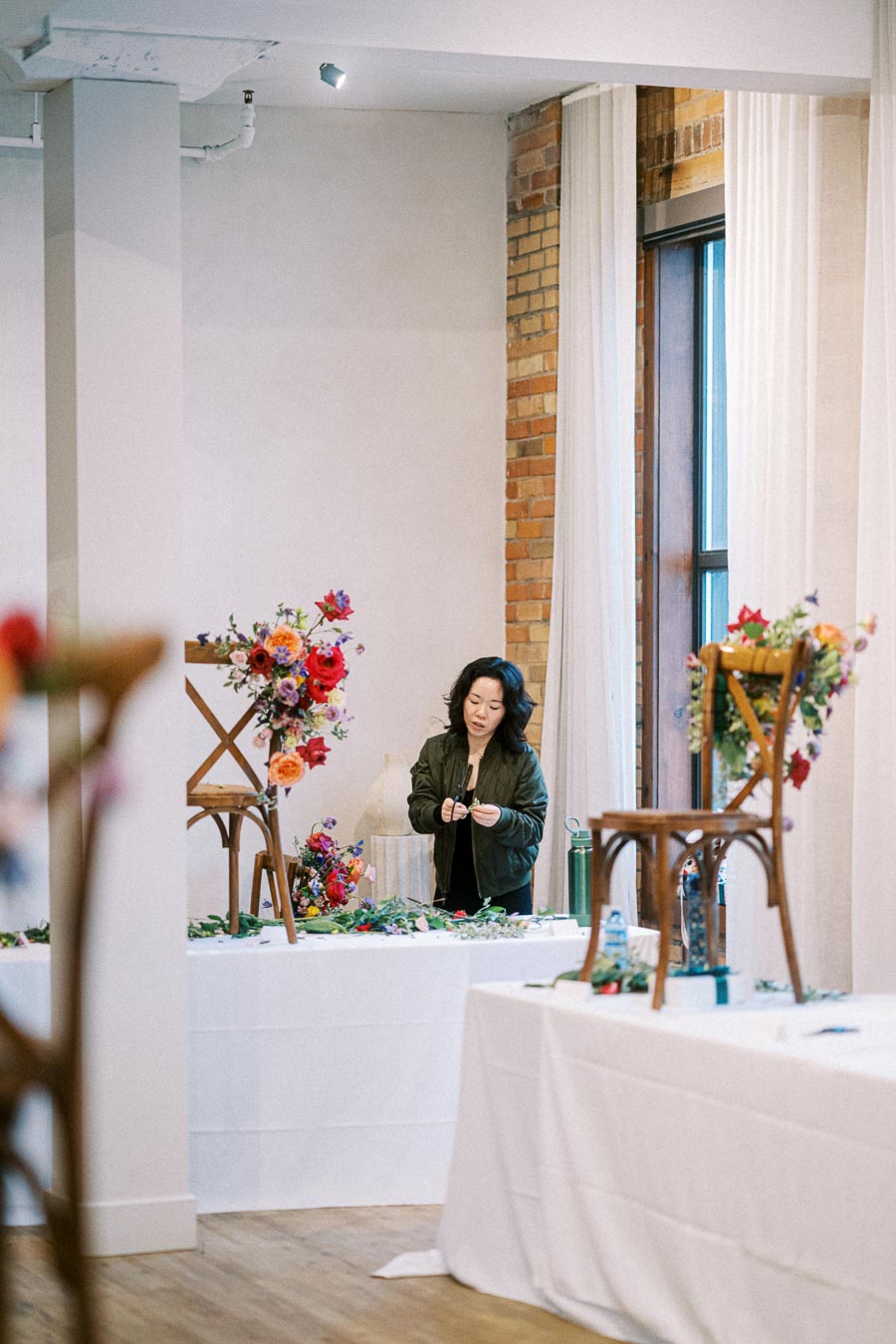 Female florist arranging colorful floral decorations on wooden chairs in a bright event space with brick and white walls.