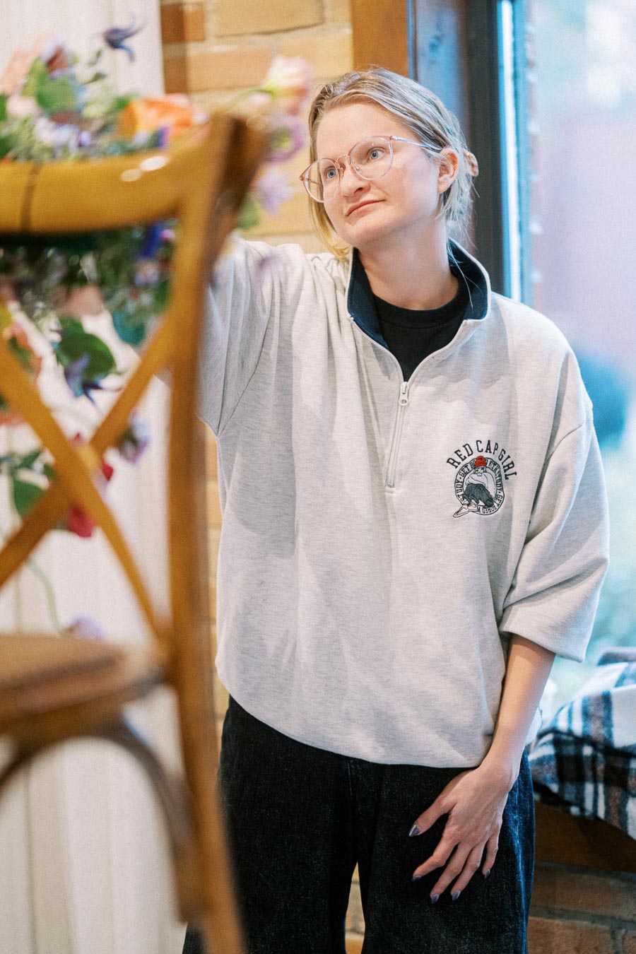 A person with glasses and a gray sweatshirt arranging flowers near a wooden chair, in a cozy, warmly lit room.