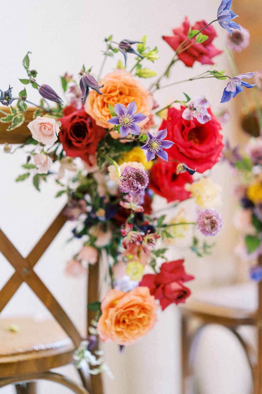 Colorful floral arrangement on a chair, featuring orange roses, red and pink roses, purple clematis, and various greenery, ideal for wedding or event decor.