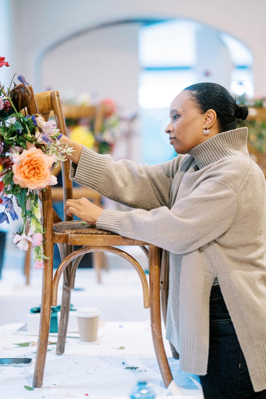 A person carefully arranging a vibrant floral display on a wooden chair, focusing intently in a well-lit indoor setting.