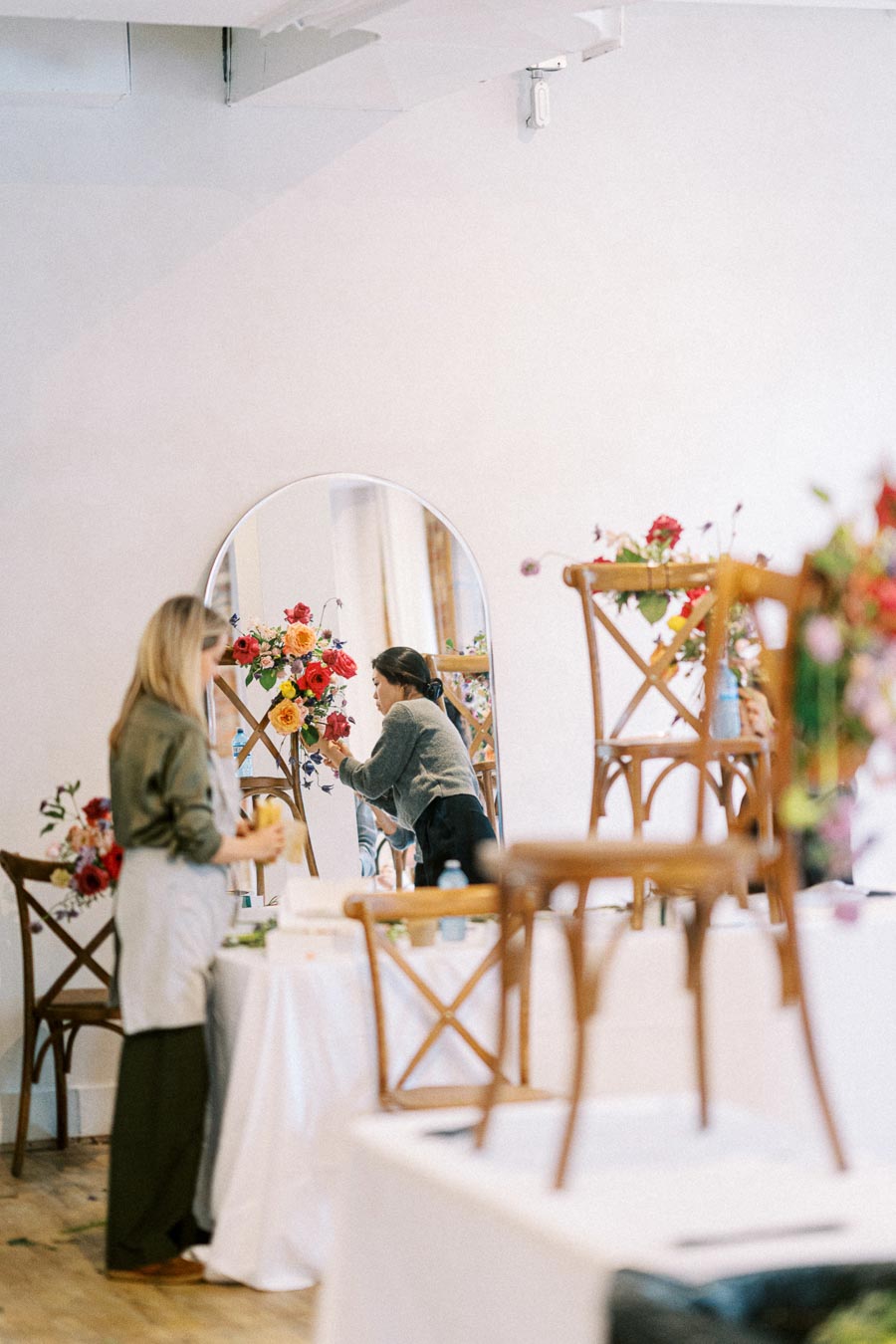 A florist arranges a vibrant floral display in front of a mirror, reflecting her creative process, with wooden chairs and more flower arrangements in a bright, airy room.