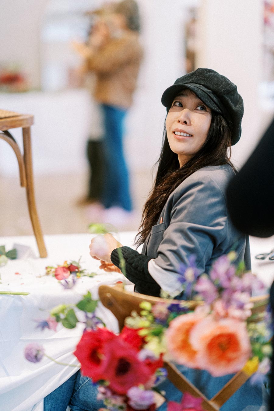 A woman wearing a black hat sits at a table arranged with colorful flowers, smiling and looking upwards in a bright indoor setting.