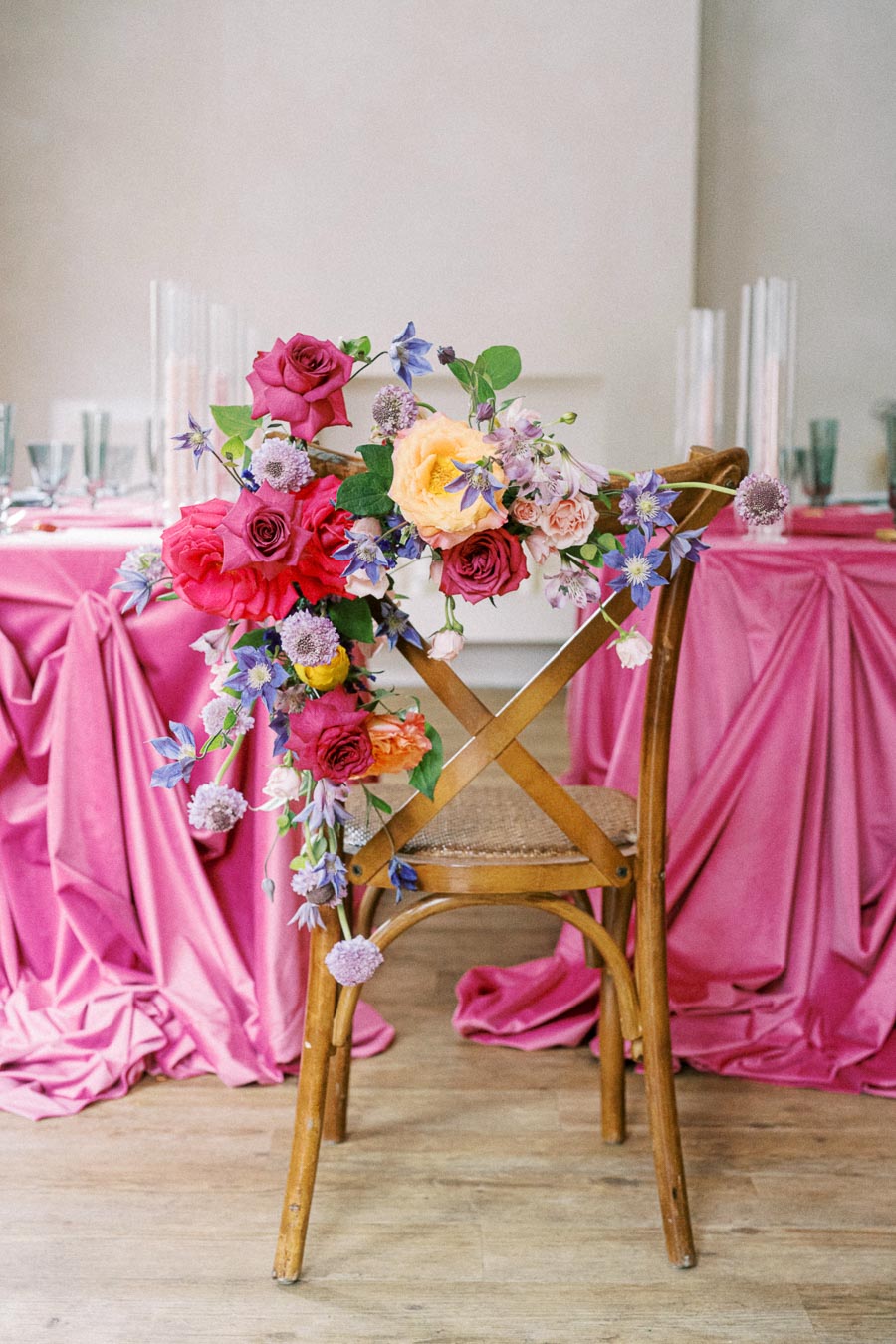 Elegant wooden chair decorated with vibrant floral arrangement featuring pink, yellow, and purple flowers, set against a table with draped pink tablecloth, perfect for a romantic wedding or special event setup.