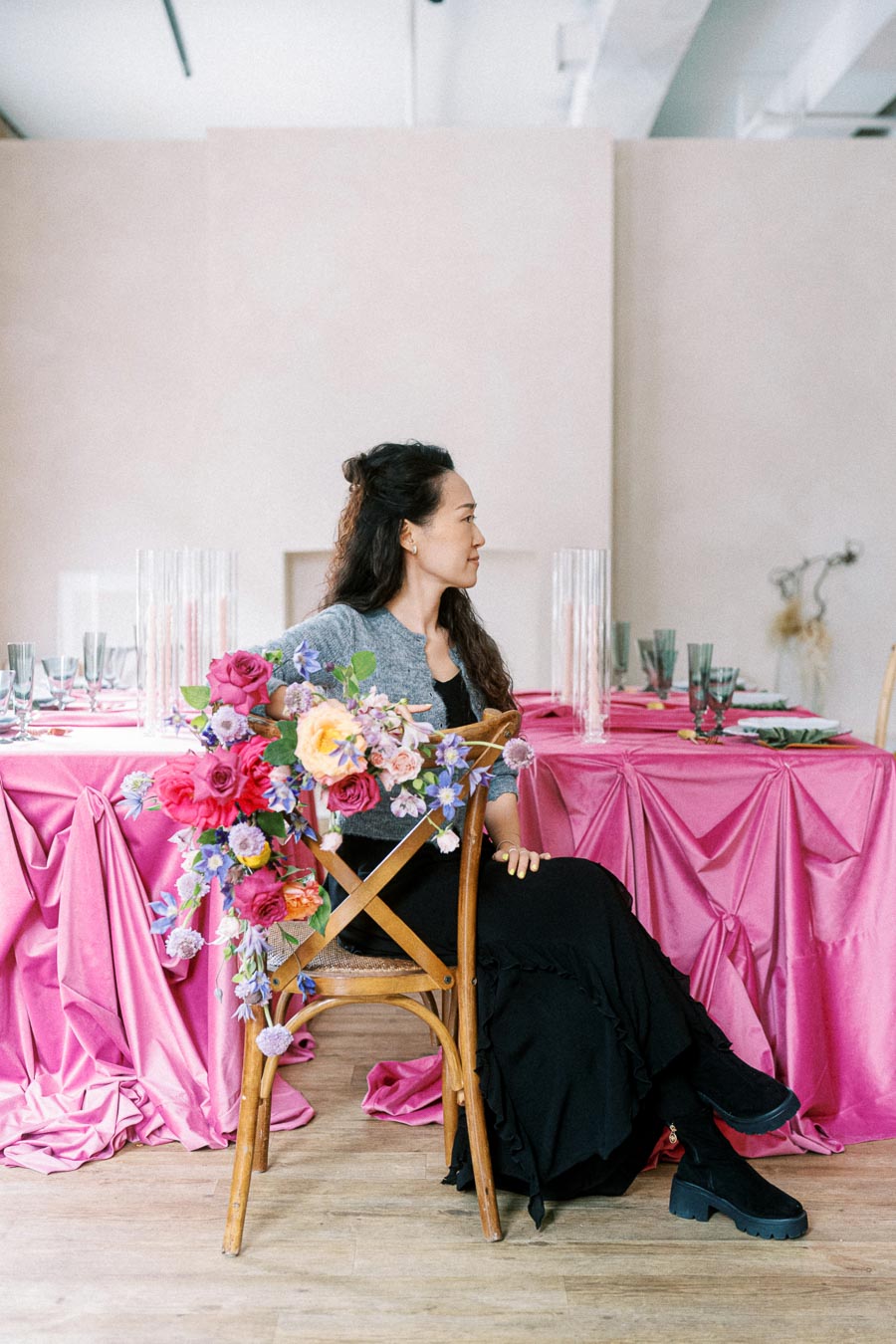 A woman sitting on a wooden chair, adorned with a vibrant floral arrangement, next to a table with a pink tablecloth and elegant glassware.