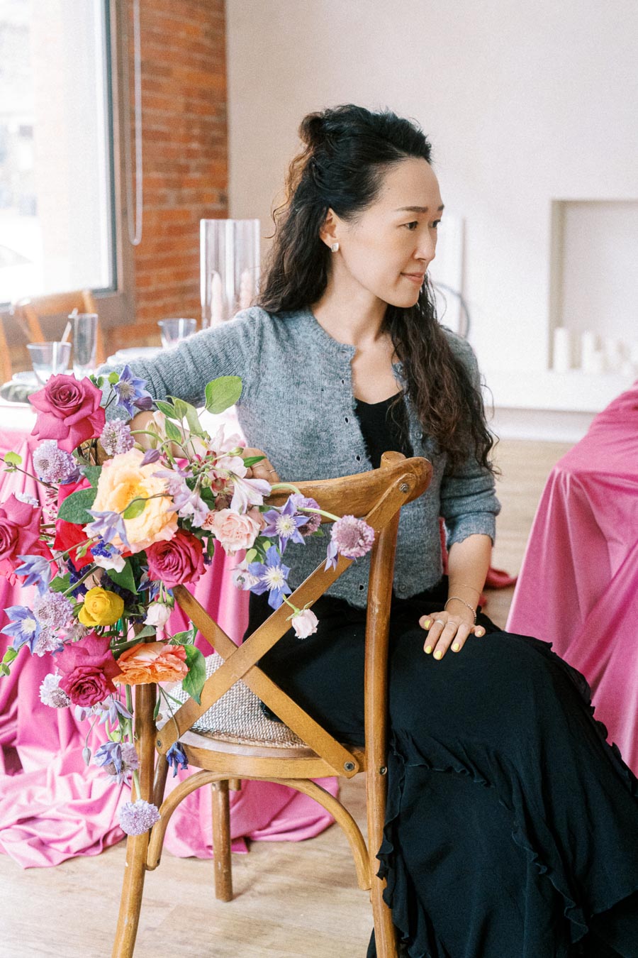 A woman with long dark hair sits on a wooden chair adorned with a colorful floral arrangement, featuring roses and various blooms, against a backdrop of pink fabric in a cozy room.