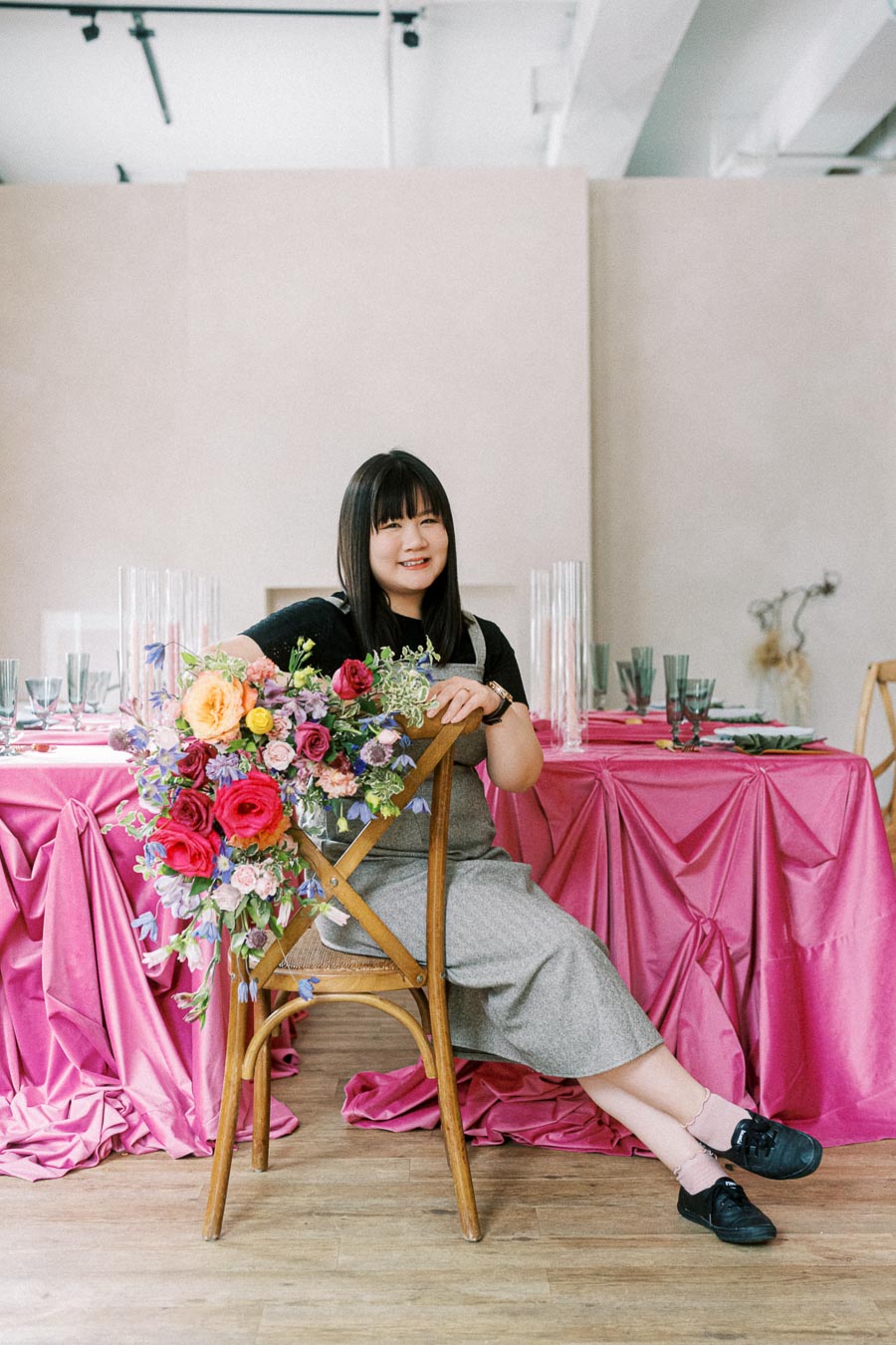 A woman seated on a wooden chair adorned with a vibrant bouquet of pink, purple, and yellow flowers, positioned in front of a table draped in bright pink cloth with elegant table settings, showcasing a modern event or wedding setup.